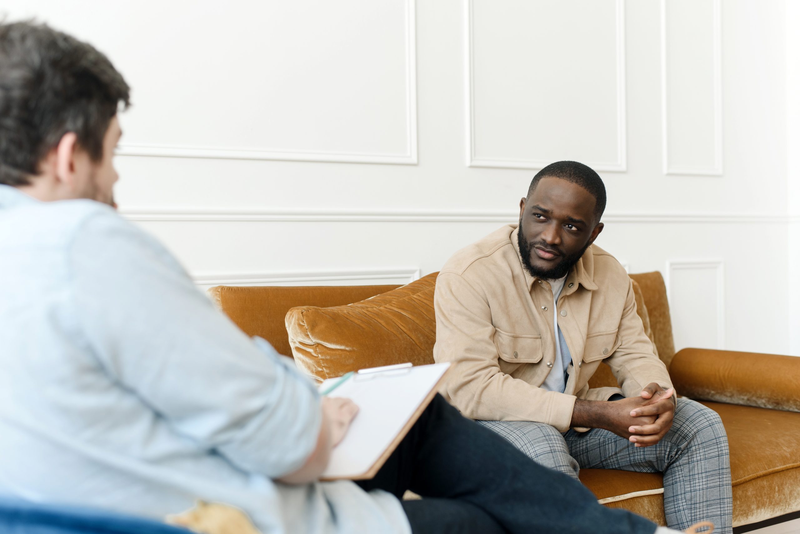 An irritated African American emotionally talks about problems to a psychologist at a session, a confused unhappy young man undergoes therapy with a psychiatrist.